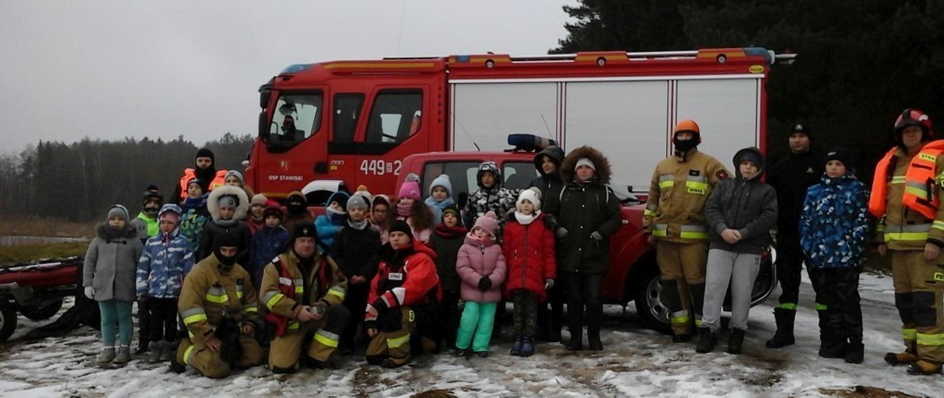 Na zdjęciu widać dzieci ze strażakami, w tyle pojazdy strażackie.