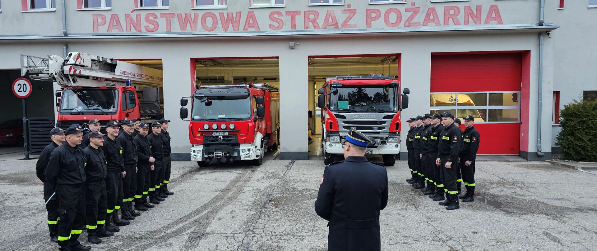 Strażacka zbiórka na utwardzonym placu. Po lewej i prawej stronie stoją w dwuszeregach strażacy w czarnych mundurach. W tle czerwone pojazdy pożarnicze i budynek garażu z napisem Państwowa Straż Pożarna.