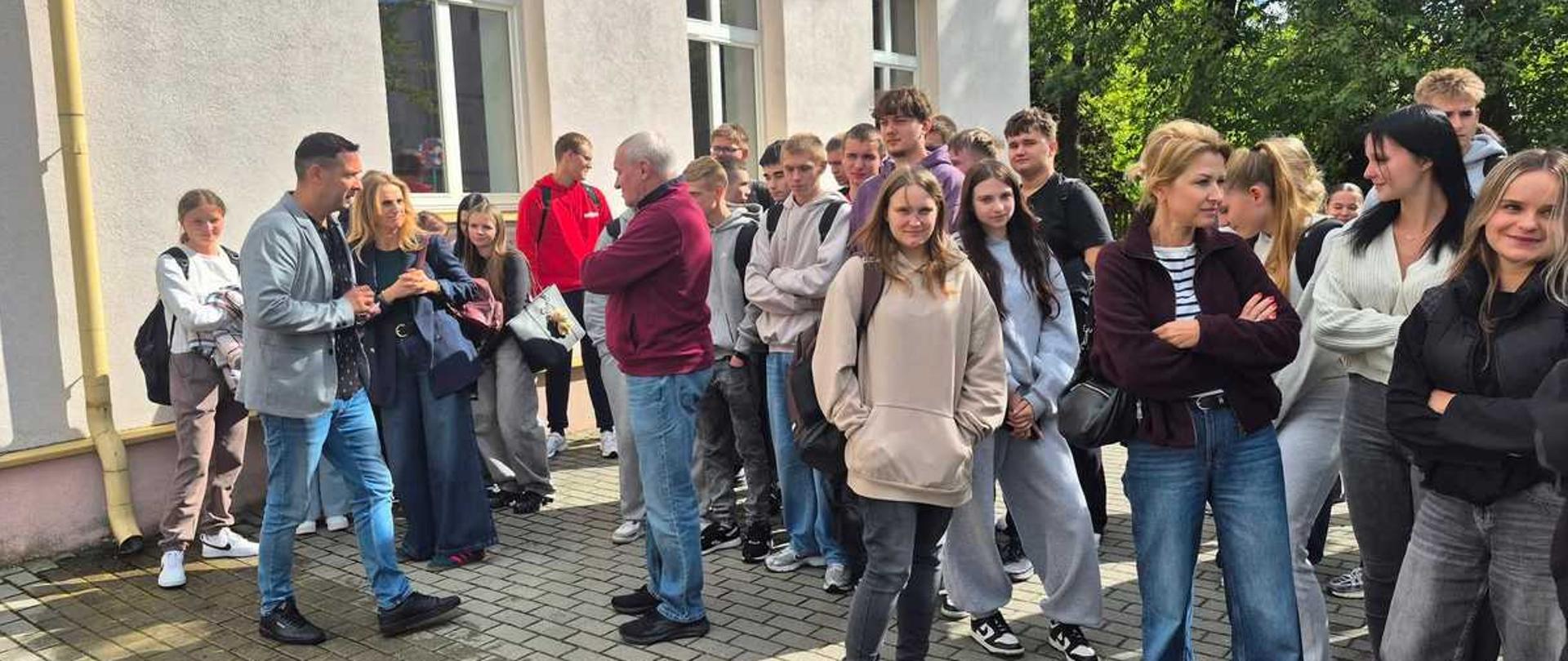 "Zdjęcie przedstawia dużą grupę młodzieży oraz kilkoro dorosłych stojących na zewnątrz, przed jasnym budynkiem w słoneczny dzień.
Sceneria i tło
W tle po lewej stronie widoczny jest fragment piętrowego, klasycznego budynku o jasnotynkowanych ścianach z dużymi, prostokątnymi oknami w białych ramach. Po prawej stronie, za zgromadzonymi ludźmi, rosną gęste, zielone drzewa, przez które przebija się słońce. Wszyscy stoją na chodniku wyłożonym szarą kostką brukową. Światło słoneczne rzuca wyraźne cienie na ziemię i elewację budynku.
Osoby na zdjęciu
Na zdjęciu znajduje się około dwudziestu osób, w większości w wieku szkolnym (nastolatkowie), ubranych w luźne, codzienne stroje sportowe i casualowe – bluzy z kapturem, jeansy, dresy oraz sneakersy. Wśród nich wyróżnia się kilka osób stojących na pierwszym planie:
• Po lewej stronie: Mężczyzna w średnim wieku w jasnoszarej marynarce, wzorzystej koszuli i niebieskich jeansach rozmawia z kobietą w ciemnej, eleganckiej marynarce i szerokich spodniach. Obok nich stoi młoda dziewczyna z plecakiem.
• W centrum: Starszy, nieco tęgi mężczyzna z siwiejącymi włosami, ubrany w bordową bluzę i jeansy, stoi bokiem z założonymi rękami i rozmawia z otaczającą go młodzieżą. Tuż przed nim stoi dziewczyna w jasnobeżowej bluzie z kapturem i ciemnych spodniach, patrząca w stronę obiektywu.
• Po prawej stronie: Na pierwszym planie stoi kobieta w bordowej, rozpinanej bluzie, pasiastej bluzce i klasycznych jeansach, z założonymi rękami. Obok niej i za nią tłoczą się kolejne uśmiechnięte dziewczęta i chłopcy, z których część patrzy bezpośrednio w stronę aparatu."