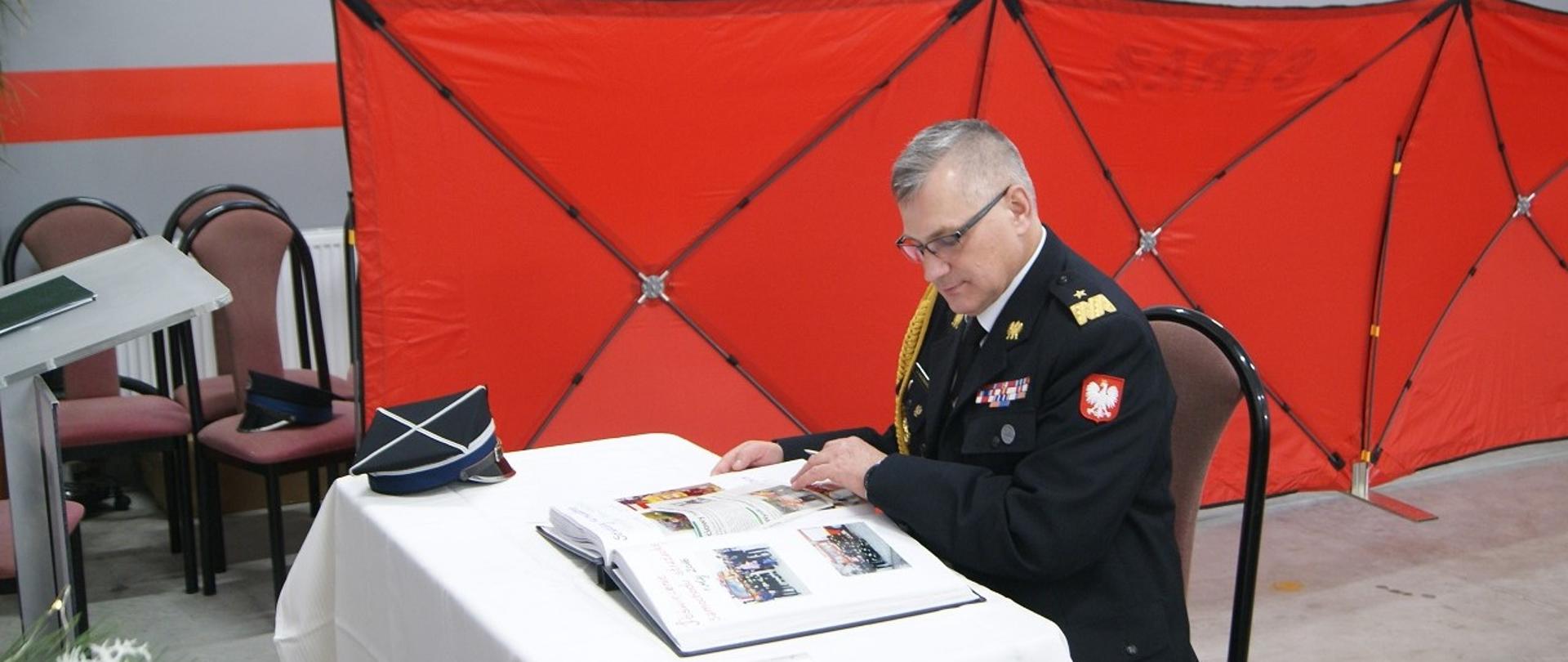 Komendant wojewódzki, generał, dokonuje wpisu w księdze pamiątkowej jednostki OSP. Na stole kronika, czapka, w tle parawan oddzielający wyposażenie remizy od miejsca uroczystości.