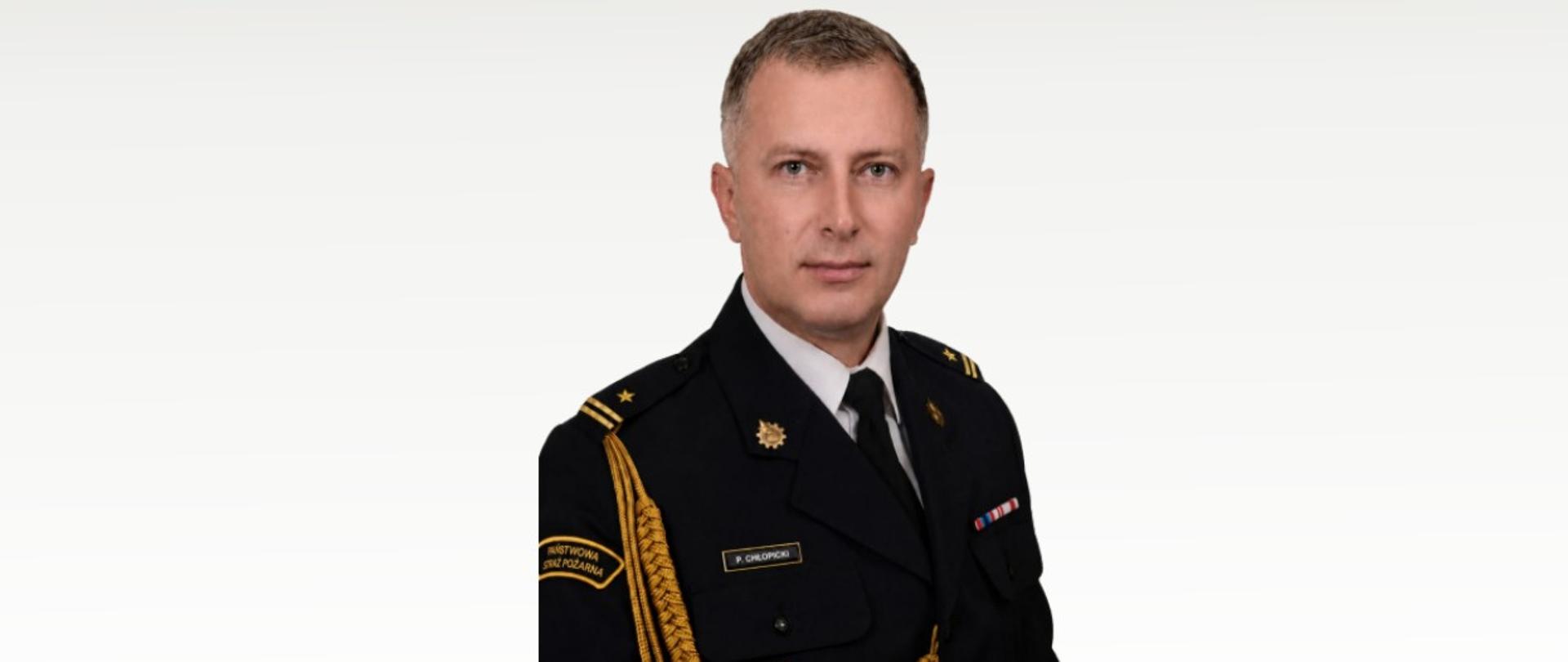 Zdjęcie przedstawia zdjęcie Zastępcy Komendanta Miejskiego Państwowej Straży Pożarnej mł. bryg. Piotra Chłopickiego ubranego w mundur galowy. Zdjęcie wykonane na białym tle.