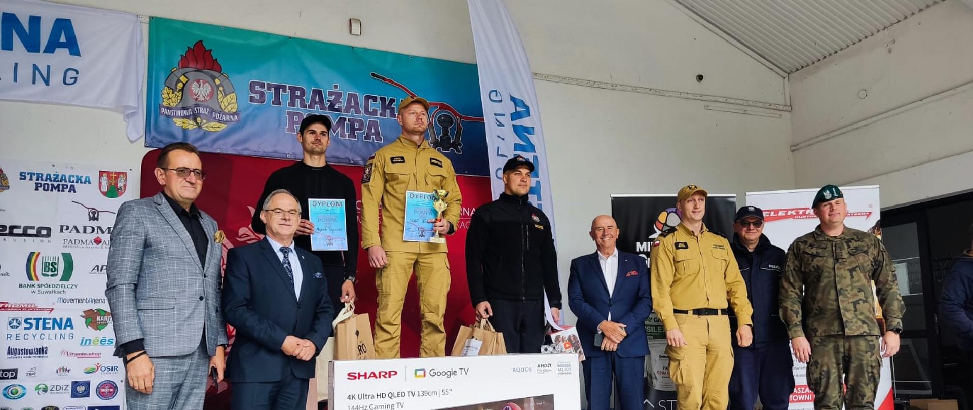 Grupa osób na podium z nagrodami podczas zawodów strażackich.