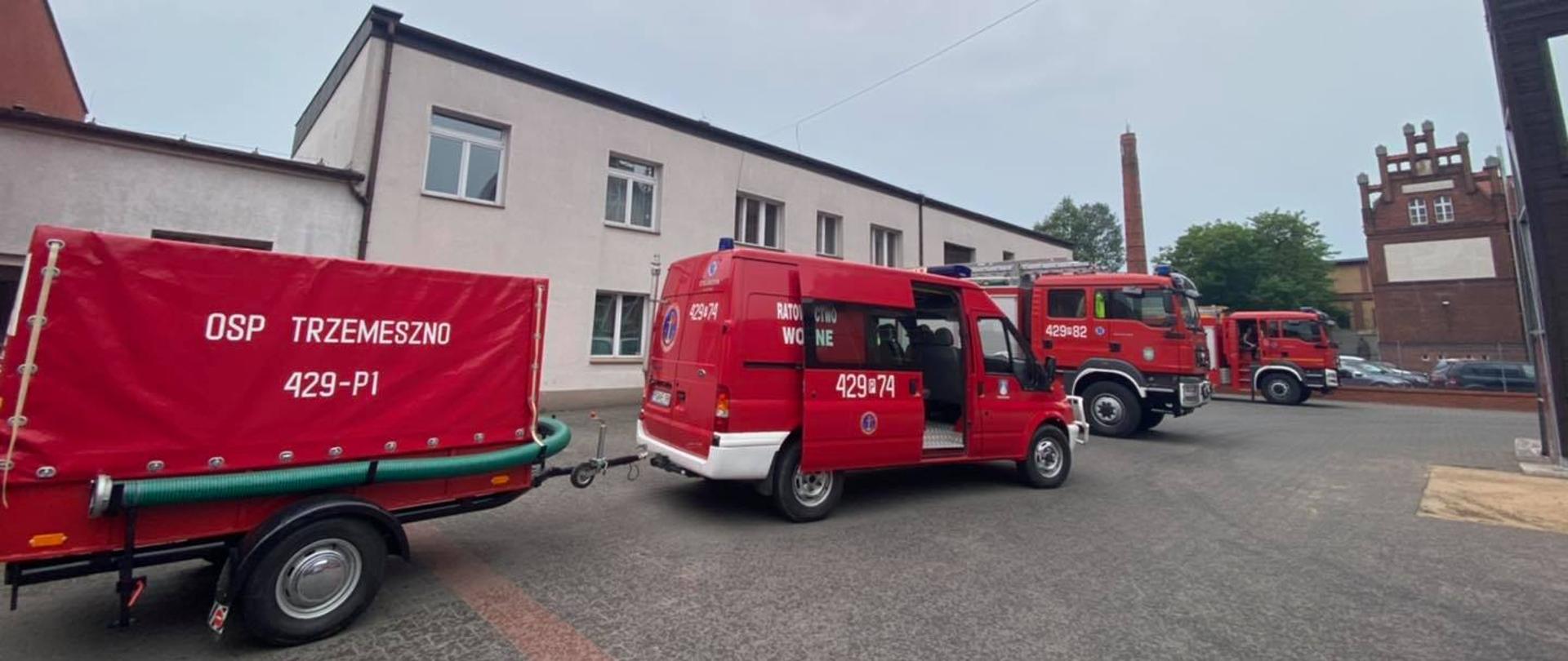 Pochmurno. Czerwony wozy strażackie stoją na placu KP PSP w Gnieźnie. W tle biały budynek komendy oraz czarna wspinalnia.
