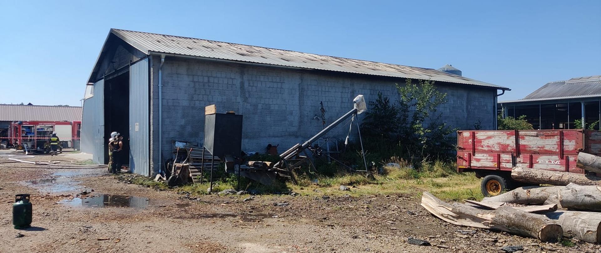 Hala produkcyjna z widocznymi śladami pożaru. Pod halą stoi dwóch strażaków. W tle samochody strażackie. Przed halą przyczepa, bele drewna oraz maszyny.