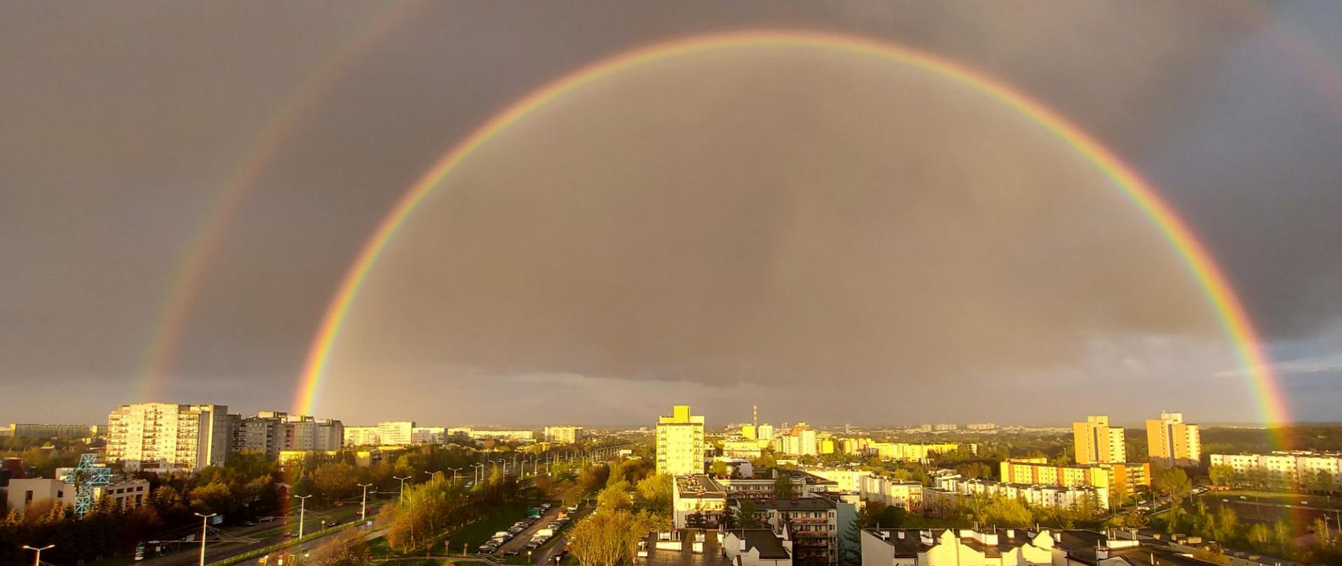 Kolorowe zdjęcie - widziane z góry bloki mieszkalne i ulice. Nad nimi rozciąga się półkolista, podwójna tęcza, która zajmuje dwie trzecie kadru, wchodząc częściowo na budynki
