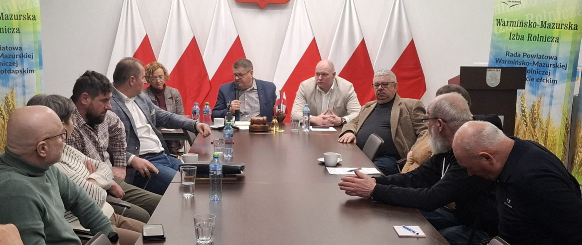 Duży stół, na którym leżą dokumenty oraz stoją szklanki i butelki z wodą. Wokół stołu siedzą ludzie i rozmawiają. 