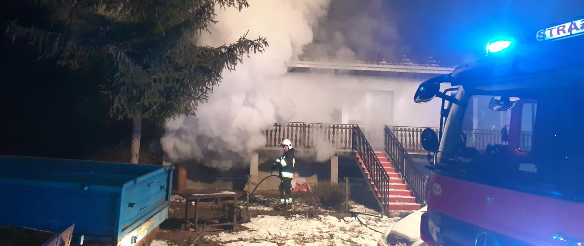 Zdjęcie wykonane w porze nocnej obrazuje częściowo budynek mieszkalny, z którego przez okna piwniczne wydostają się duże kłęby jasnego dymu. Centralnie na zdjęciu stoi strażak OSP. Z prawej strony widoczny jest przód samochodu strażackiego , a z lewej część przyczepy, drzewo i krzewy rosnące obok budynku.