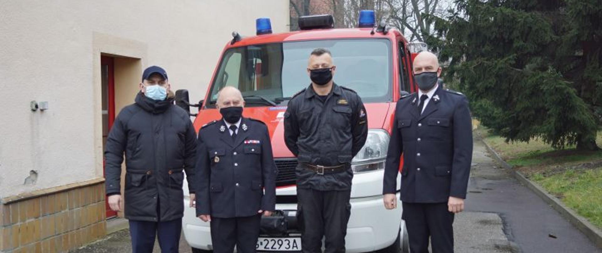 Czterej mężczyźni stojący obok siebie przed czerwonym pojazdem pożarniczym widzianym z przodu. Dwaj z nich ubrani są w czarne, galowe mundury strażackie. Jeden w czarny mundur dowódczo – sztabowy, a ostatni w ciemną kurtkę i niebieskie spodnie. W tle po lewej ściana budynku, a po prawej zielony świerk rosnący na trawniku. 