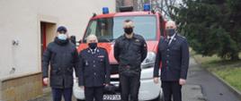 Czterej mężczyźni stojący obok siebie przed czerwonym pojazdem pożarniczym widzianym z przodu. Dwaj z nich ubrani są w czarne, galowe mundury strażackie. Jeden w czarny mundur dowódczo – sztabowy, a ostatni w ciemną kurtkę i niebieskie spodnie. W tle po lewej ściana budynku, a po prawej zielony świerk rosnący na trawniku. 