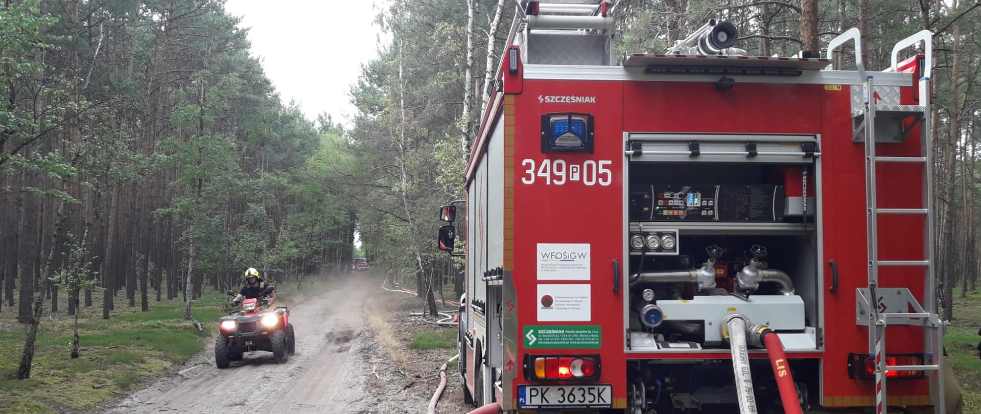 Na zdjęciu widać po lewej stronie samochód gaśniczy w kolorze czerwonym, posiada sygnalizację ostrzegawczą oraz numery operacyjne. Do samochodu podłączone są linie wężowe, które tworzą magistralę wodną. Po lewej stronie jedzie quad w kolorze czerwonym, na włączonych światłach. Widać dukt leśny. Wokół drzewa, korony zielone. Niebo zachmurzone.