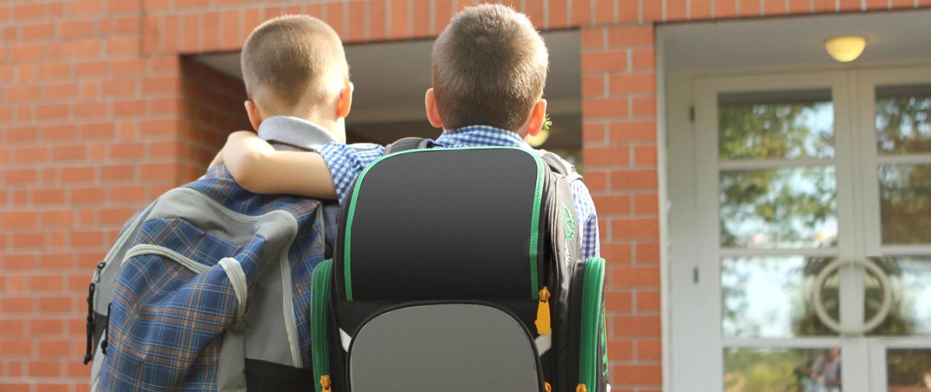 Zdjęcie przedstawia dwóch chłopaków przytulających się do siebie kierujących w stronę szkoły. Na plecach mają założone plecaki.