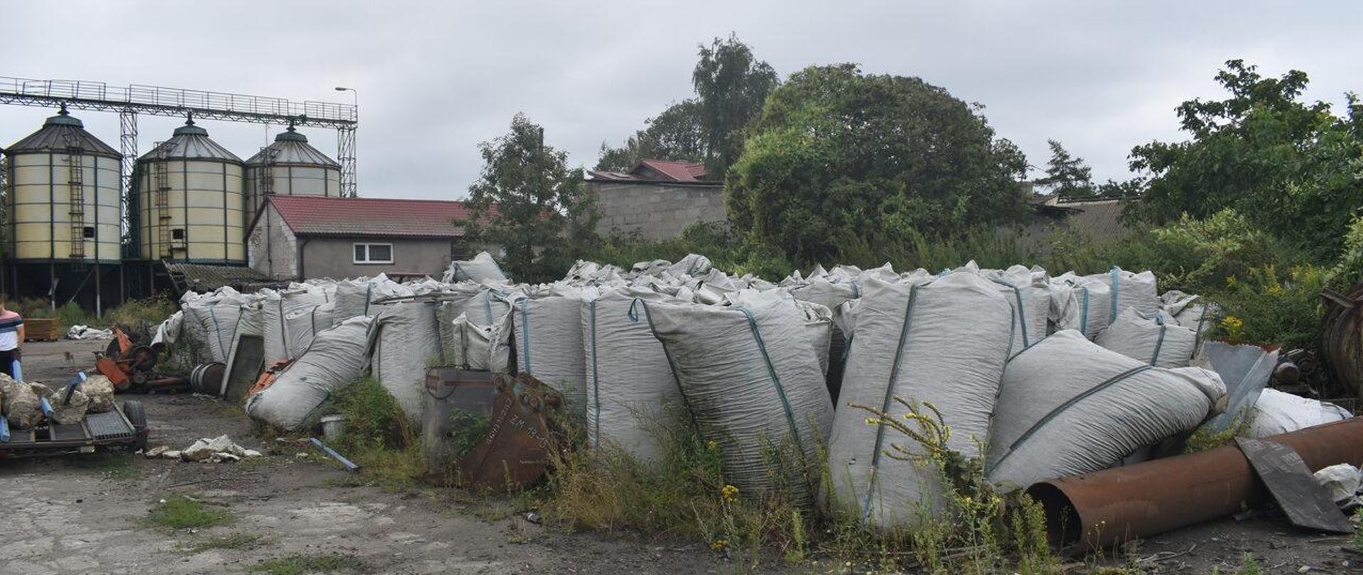 Kilkadziesiąt białych worków typu big-bag stoi obok siebie na nieutwardzonym podłożu. W tle widać zabudowanie gospodarskie skryte częściowo za drzewami oraz trzy stalowe silosy.