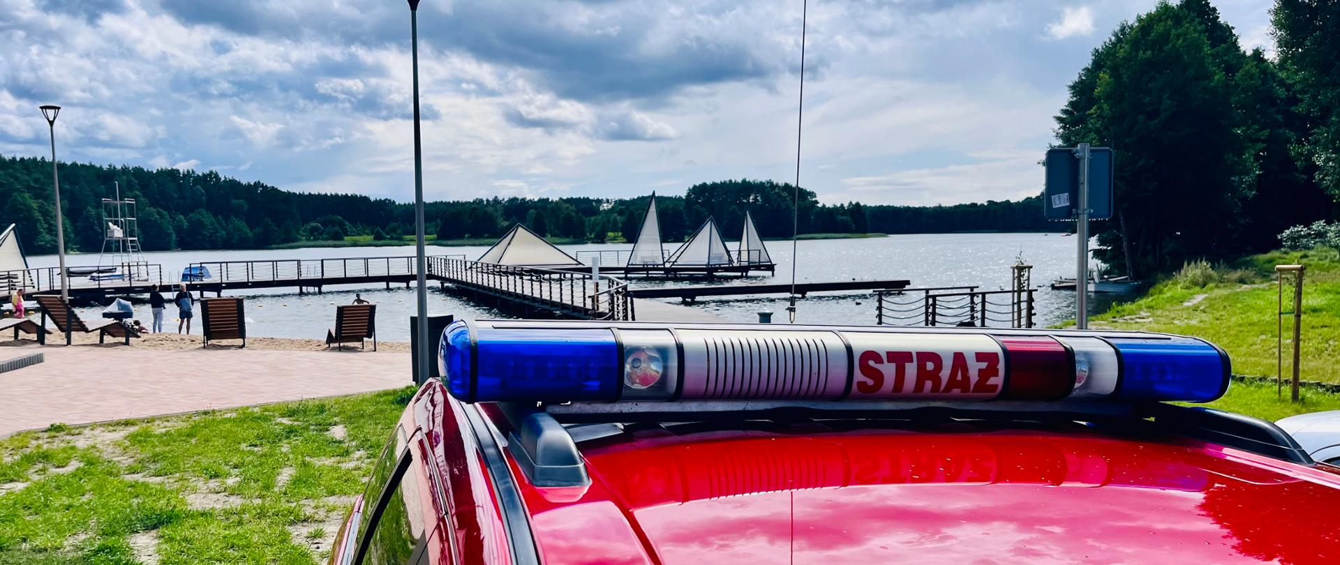 Na zdjęciu widać dach samochodu pożarniczego z sygnałami świetlnymi, w oddali jezioro i pomosty.