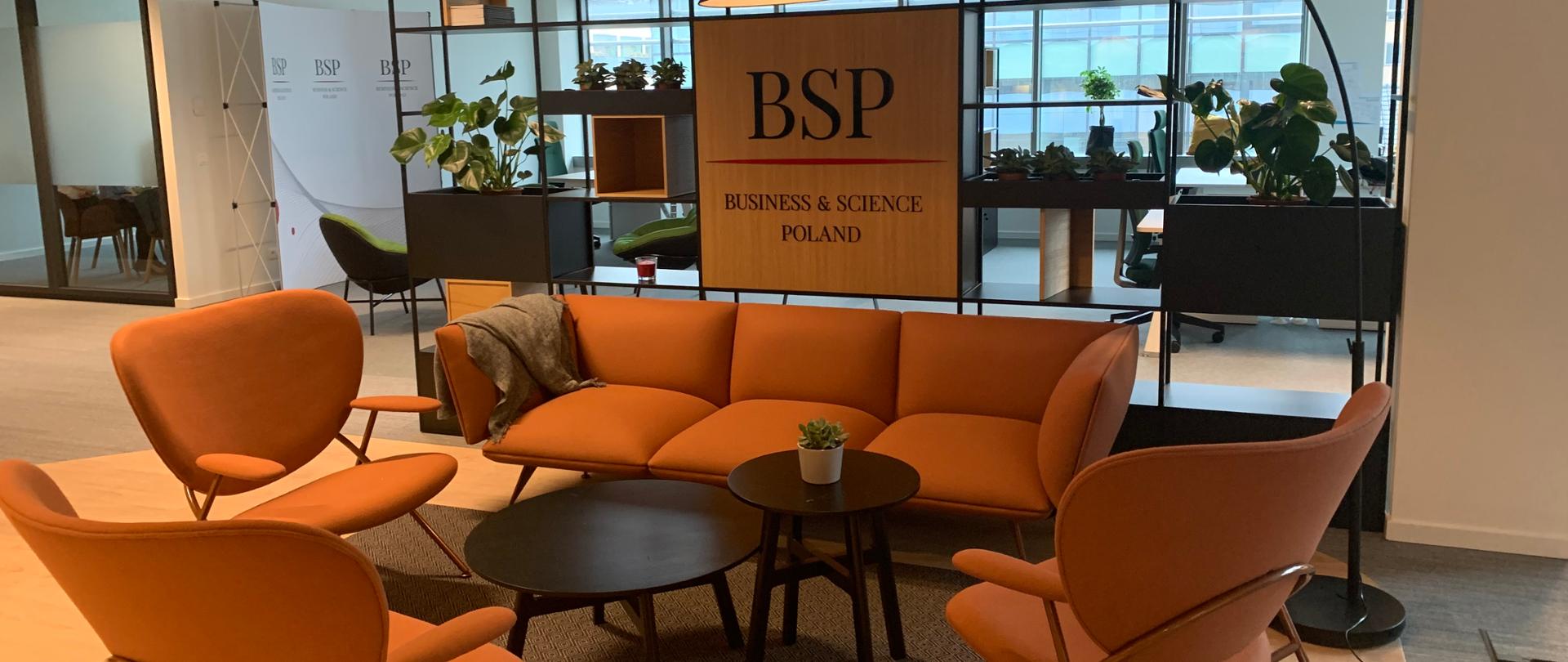 Interior of the office in Brussels. Two wooden round tables next to each other, 3 armchairs around and one orange sofa. On one table there is a flower in a pot. from the Back Bunk shelves with flowers. In the middle a banner with the word "BSP" underlined with a red line. Below, the inscription "Business & science Poland". The rest of the office is visible in the background.