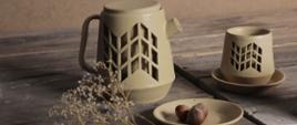 Zdjęcie: na deskach ceramiczny dzbanek, filiżanka i talerzyk na którym leżą trzy orzechy laskowe.