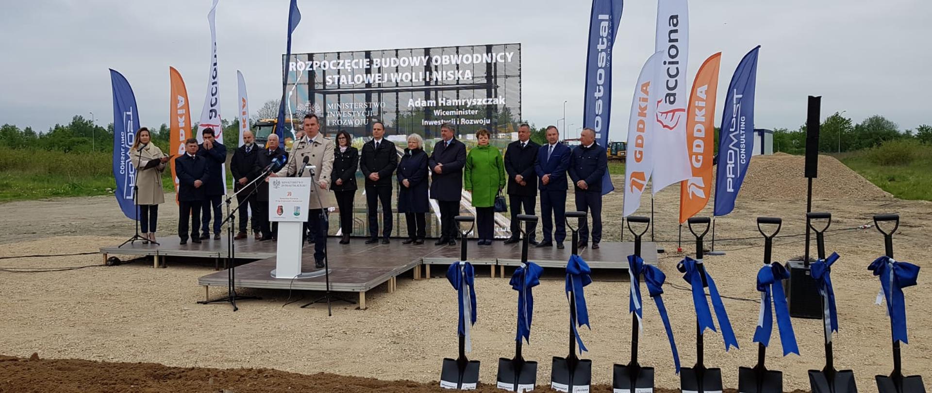 Na scenie w terenie otwartym stoi grupa ludzi. Za nimi baner z napisem: Rozpoczęcie budowy obwodnicy Stalowej Woli i Niska. Na pierwszym planie w mównicy przemawia wiceminister Adam Hamryszczak.