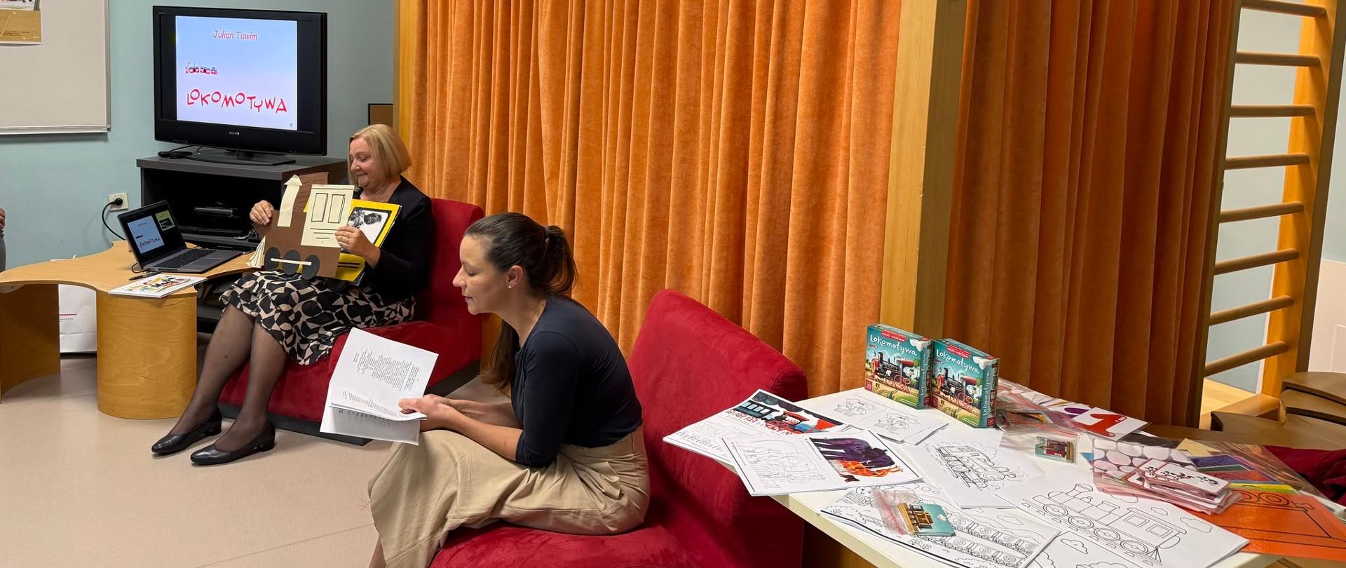 Dwie prowadzące przygotowują materiały do spotkania z dziećmi. Na ekranie widoczny jest tytuł Lokomotywa Juliana Tuwima, a na stole leżą kolorowanki, ilustracje pociągów i materiały do zajęć.