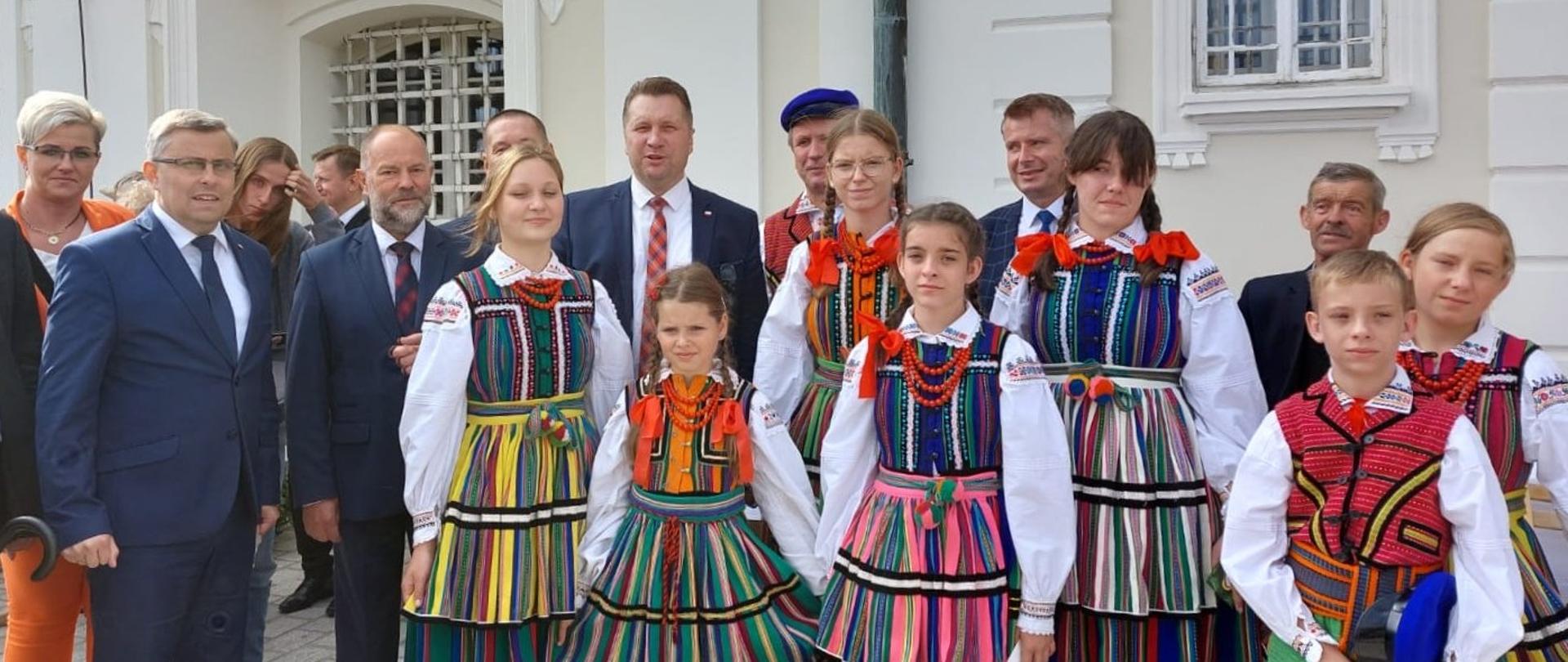 Grupa dzieci w ludowych strojach, za nimi minister Czarnek w otoczeniu ludzi w garniturach.