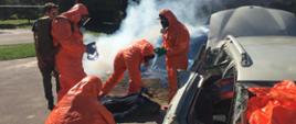 Czterech uczestników szkolenia w ochronnych kombinezonach i maskach sprawdza stan ofiary wypadku leżącej obok rozbitego samochodu