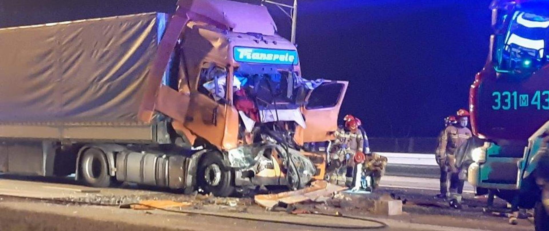 Zdjęcie wykonane w porze nocne. Na zdjęciu widać rozbity samochód ciężarowy. Do pojazdu podpięta jest naczepa. Kabina kierowcy jest zniszczona, na ziemi leżą elementy pojazdu oraz wyposażenia kabiny. Lewe drzwi prowadzące do kabiny, od strony kierowcy są otwarte. Przed pojazdem stoją strażacy ubrani w żółte ubrania specjalne z elementami odblaskowymi. Na głowach mają czerwone hełmy. Obok nich znajduje się również pojazd pożarniczy w kolorze czerwonym z zapalonymi niebieskimi światłami alarmowymi. 