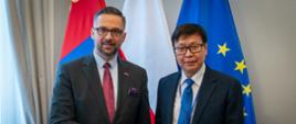 Minister Kulasek stoi obok mężczyzny w garniturze, podają sobie ręce, za nimi pod białą ścianą flagi Polski, UE i Mongolii.