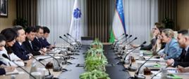 Sala konferencyjna, w której siedzą dwie grupy osób naprzeciwko siebie przy długim stole. Na stole rozmieszczone są mikrofony i rośliny. W tle widoczne są dwie flagi, w tym flaga Uzbekistanu, oraz biała flaga z logiem i tekstem. Nad stołem wiszą okrągłe lampy.