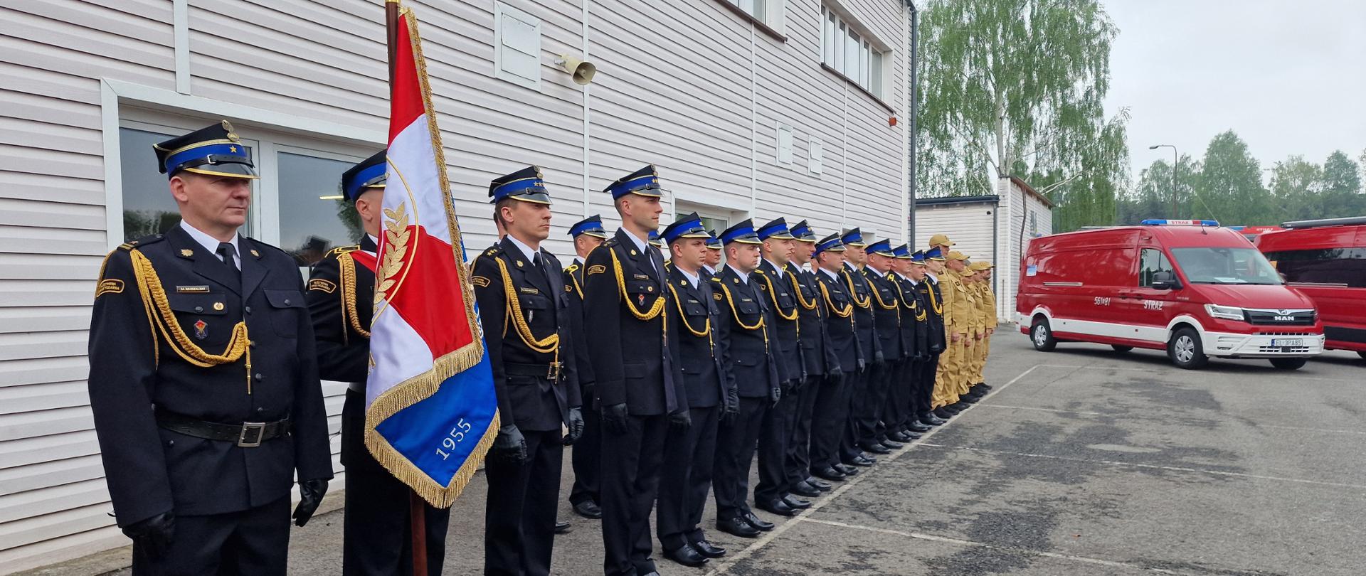 Na zdjęciu przedstawiono strażaków ustawionych w szeregu podczas oficjalnej uroczystości przed budynkiem straży pożarnej. Większość funkcjonariuszy ma na sobie galowe granatowe mundury z czapkami i ozdobnymi sznurami, a w dalszej części szeregu stoją strażacy w jasnych mundurach służbowych. Na pierwszym planie widoczny jest poczet sztandarowy trzymający sztandar z biało-czerwonymi barwami. Strażacy stoją na baczność, co wskazuje na ceremonialny charakter wydarzenia. Po prawej stronie stoją czerwone pojazdy strażackie, w tym samochód ratowniczy z niebieskimi światłami na dachu. W tle widać drzewa oraz pochmurne niebo. Całość jest uroczystym apelem organizowanym przez Państwową Straż Pożarną.