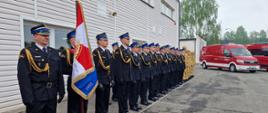 Na zdjęciu przedstawiono strażaków ustawionych w szeregu podczas oficjalnej uroczystości przed budynkiem straży pożarnej. Większość funkcjonariuszy ma na sobie galowe granatowe mundury z czapkami i ozdobnymi sznurami, a w dalszej części szeregu stoją strażacy w jasnych mundurach służbowych. Na pierwszym planie widoczny jest poczet sztandarowy trzymający sztandar z biało-czerwonymi barwami. Strażacy stoją na baczność, co wskazuje na ceremonialny charakter wydarzenia. Po prawej stronie stoją czerwone pojazdy strażackie, w tym samochód ratowniczy z niebieskimi światłami na dachu. W tle widać drzewa oraz pochmurne niebo. Całość jest uroczystym apelem organizowanym przez Państwową Straż Pożarną.