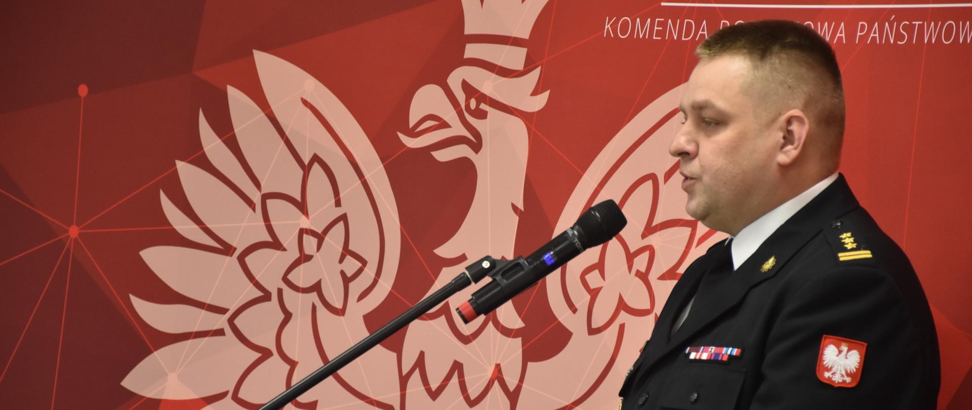 strażak stojacy przy mównicy, premawia, za nim czerowna ścianka z orłem