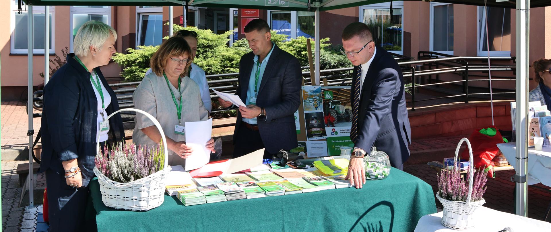 Trzy kobiety i dwóch mężczyzn stoją pod zielonym namiotem Kasy przy zielonym stoliku z rozłożonymi materiałami informacyjnymi KRUS. Stolik udekorowany jest wrzosami i cukierkami z logo KRUS.