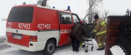 Strażak pomagający starszej osobie dotrzeć do punktu szczepień. W tle wóz strażacki OSP.