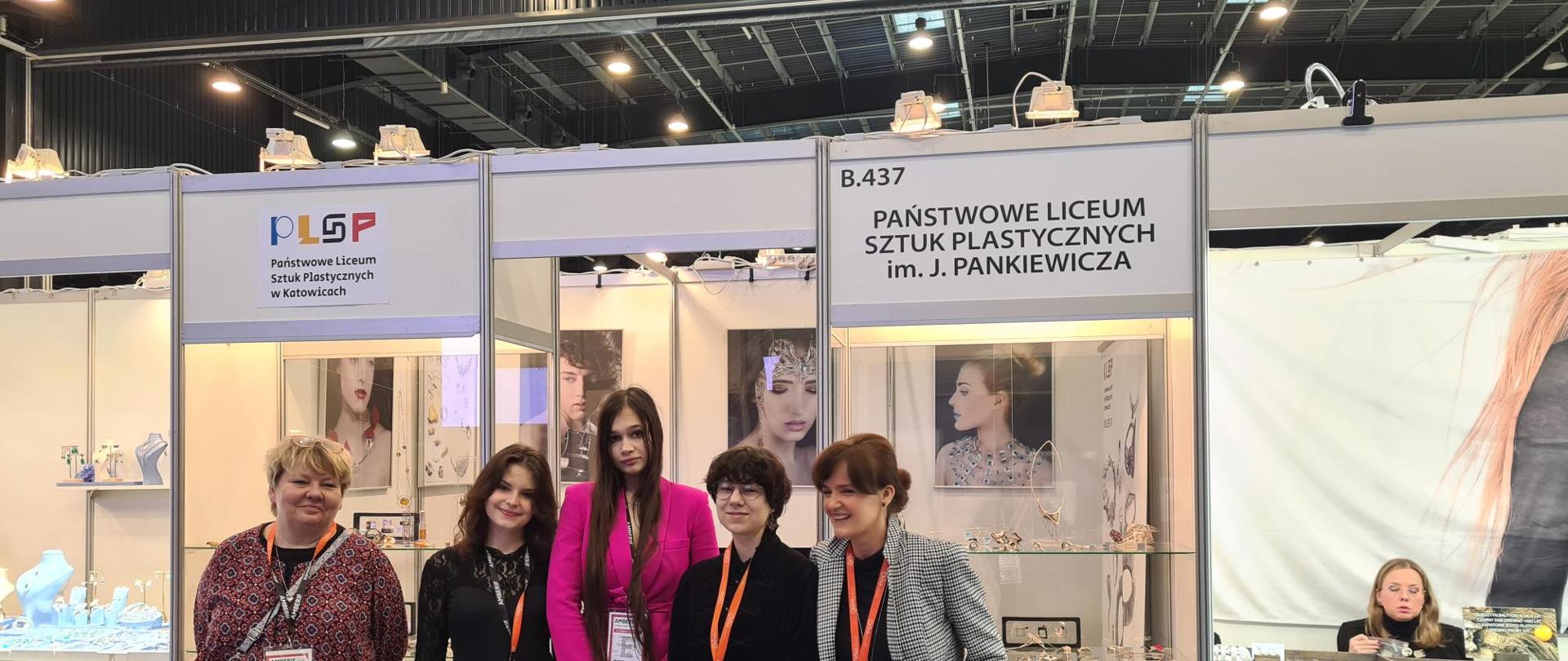 Fotografia ukazująca 5 osób z Państwowego Liceum Sztuk Plastycznych biorących udział w Międzynarodowych Targów Bursztynu & Biżuterii AmberifSPRING2024. W tle stoisko z biżuterią.