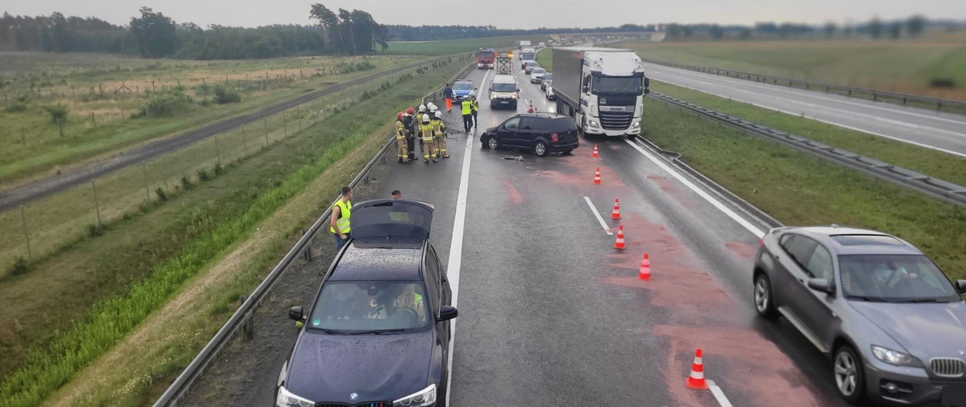 Zdjęcie zrobione z dachu samochodu pożarniczego. Na zdjęciu widać uszkodzony samochód, który stoi tyłem do kierunku jazdy. Na drodze widać sorbent oraz pachołki ostrzegawcze oddzielające teren działań