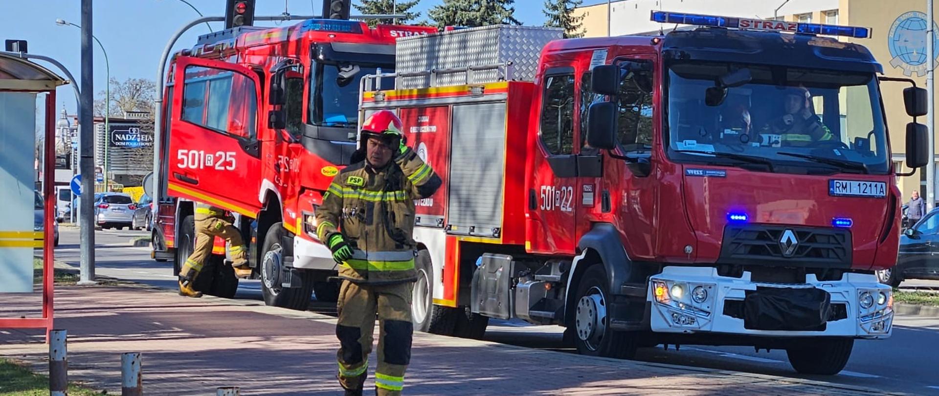 Na pierwszym planie widoczny jest strażak w pełnym umundurowaniu ochronnym koloru beżowego z żółtymi i srebrnymi pasami odblaskowymi. Ma na sobie czerwony hełm z osłoną twarzy. Strażak idzie chodnikiem w kierunku prawego dolnego rogu kadru, patrząc w stronę kamery.
Za nim, na ulicy, stoją dwa czerwone wozy strażackie. Na boku pierwszego wozu, bliżej lewej strony, widnieje biały napis "501[R]25". Przy tylnym kole tego wozu klęczy inny strażak w podobnym umundurowaniu, zajęty jakimś działaniem przy pojeździe. Drugi wóz strażacki, po prawej stronie, ma na boku napis "501[R]22" oraz tablicę rejestracyjną "RMI 1211A" z przodu. Oba wozy mają włączone niebieskie światła ostrzegawcze.
Ulica jest asfaltowa, z widocznymi pasami ruchu. Chodnik, po którym idzie strażak, jest oddzielony od jezdni niskim krawężnikiem i słupkami. W tle widać budynki miejskie o różnej architekturze i kolorystyce, drzewa (niektóre z zielonymi liśćmi, inne bez), słupy oświetleniowe oraz inne zaparkowane samochody. Niebo jest jasne, bezchmurne, co sugeruje słoneczny dzień.
Cała scena wygląda jak fragment ćwiczeń strażackich prowadzonych w terenie zabudowanym. Strażacy i wozy są przygotowani do akcji, a otoczenie wskazuje na realne warunki miejskie.
