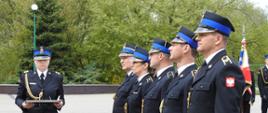 Strażacy stoją jeden obok drugiego. Na wprost stoi kobieta strażak która trzyma w rękach tacę. W tle widać zielone drzewa. Za strażakami widać sztandar. 