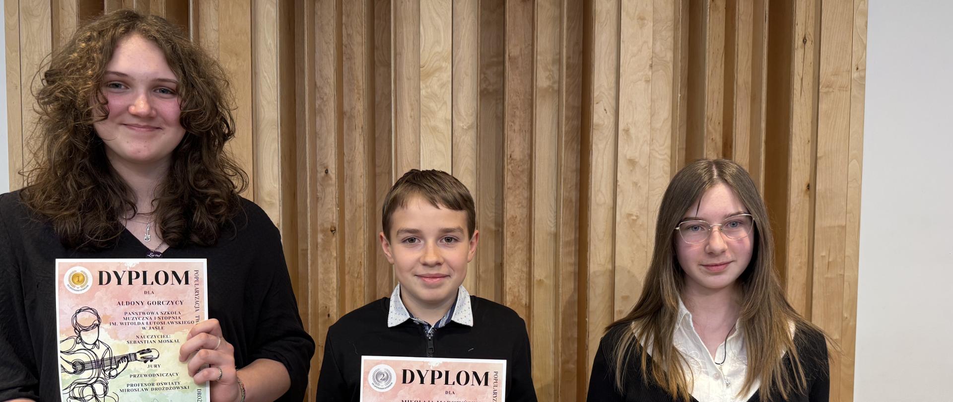 Na zdjęciu trzy osoby – dwie dziewczyny i chłopiec – stoją na tle drewnianej ściany akustycznej sali koncertowej. Wszyscy trzymają w rękach dyplomy, są elegancko ubrani w ciemne stroje z białymi akcentami. Dziewczyna po lewej ma brązowe, kręcone włosy, chłopiec w środku uśmiecha się lekko, dziewczyna po prawej ma jasne, proste włosy i okulary. Kolorystyka zdjęcia neutralna, dominują naturalne kolory drewna oraz czerń i biel strojów. 