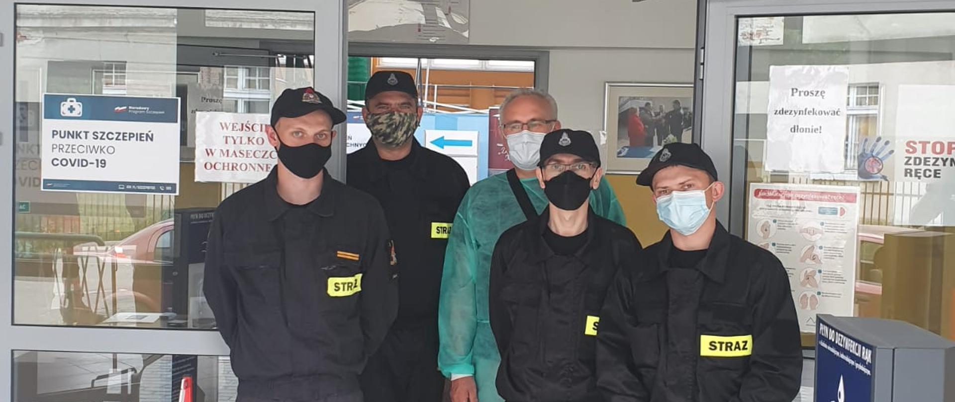 Zdjęcie przedstawia czterech umundurowanych strażaków pomagających przy punkcie szczepień. Strażakom towarzyszy jedna osoba z personelu medycznego.