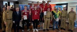 Kolorowa fotografia wykonana w dzień w pomieszczeniu. Przedstawia strażaków w strojach sportowych i służbowych stojących na podium i poza nim po wręczeniu nagród i dyplomów w XI Mistrzostwach Województwa podkarpackiego strażaków PSP w pływaniu.