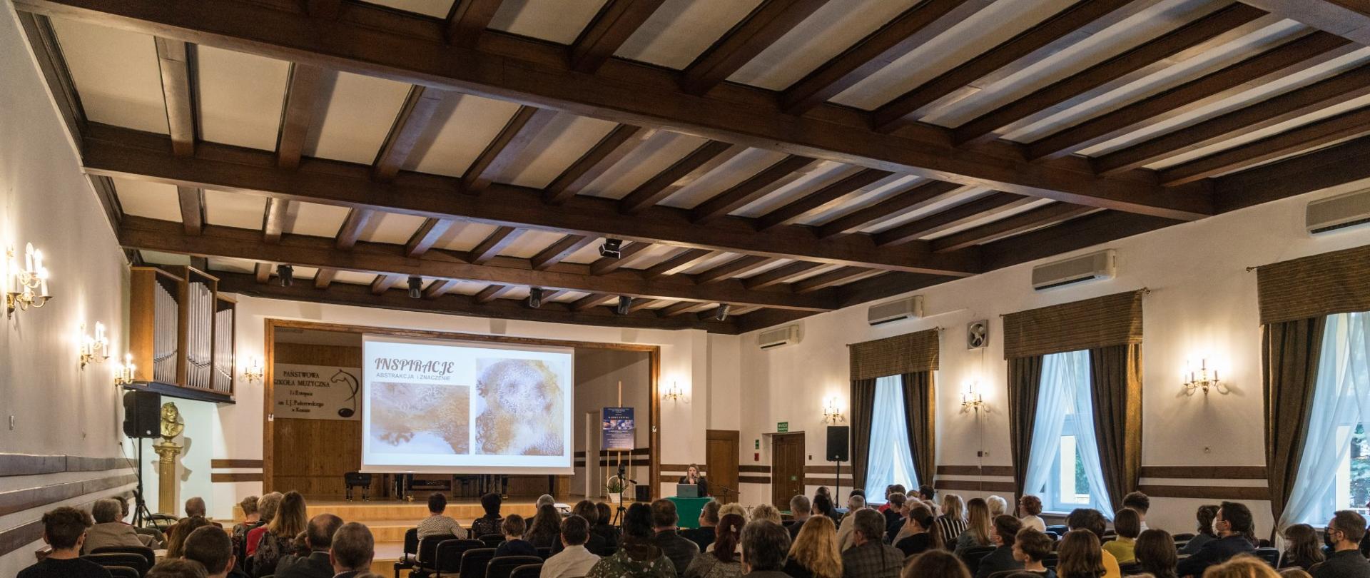 Sala koncertowa PSM Konin, koncert Barwy sztuki, na pierwszym planie publiczność, w tle scena z wyświetlaną prezentacją, 