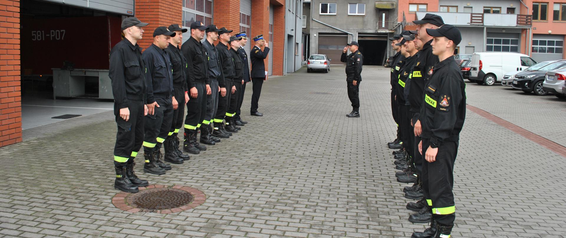17 strażaków na zmianie służby , na tle budynku Komendy, jeden melduje Komendantowi, na drugim planie samochody osobowe