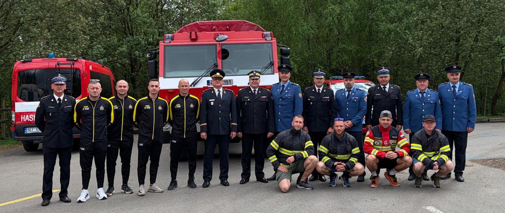 Grupowe zdjęcie siedemnastu mężczyzn pozujących przed trzema czerwonymi pojazdami strażackimi na asfaltowym placu otoczonym gęstymi, zielonymi drzewami.