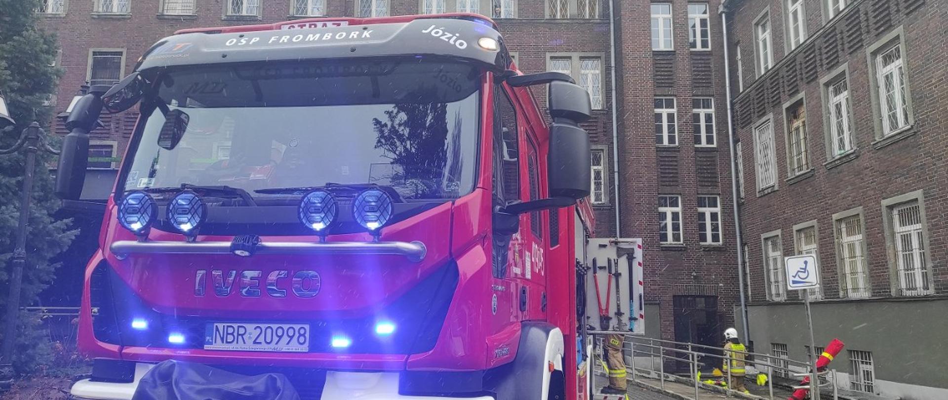 Czerwony samochód strażacki z włączonymi światłami niebieskimi na tle budynku szpitala