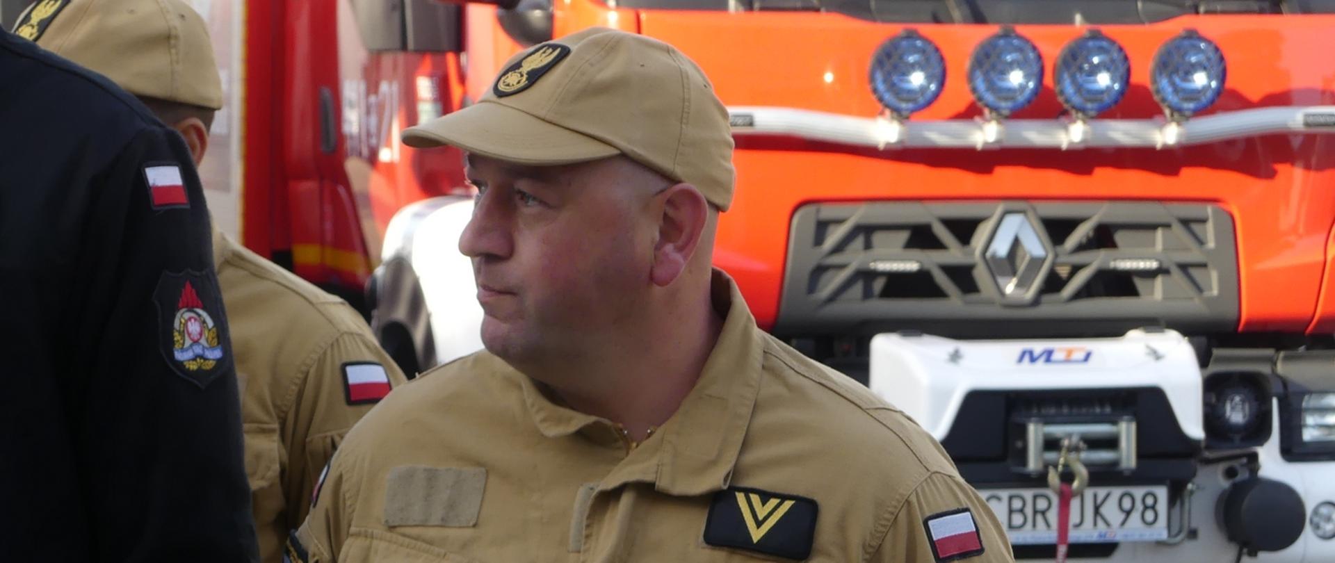 Zdjęcie przedstawia starszego ogniomistrza Jakuba Pradziada podczas uroczystej zbiorki z okazji jego przejścia na emeryturę. Strażak stoi w mundurze służbowym spoglądając w prawo w kierunku komendanta. Za nim stoi woź gaśniczy straży pożarnej, który jako kierowca często prowadził do działań ratowniczych.