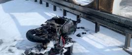 Na zdjęciu widoczny jest spalony wrak motocykla leżący na pasie awaryjnym drogi ekspresowej. Z prawej strony uszkodzone w wyniku oddziaływania ognia ekrany akustyczne. Wokół pojazdu rozlana piana gaśnicza. Na drodze lokalnej za ekranami widoczny strażak uczestniczący w akcji gaśniczej.