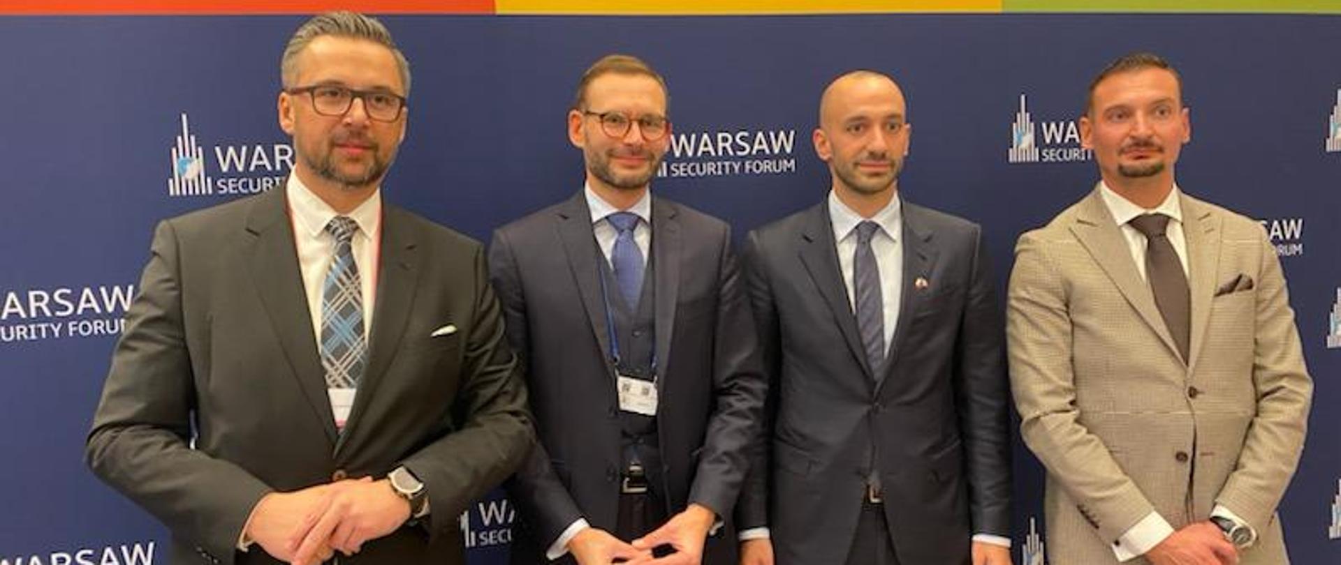 Wiceminister Marcin Kulasek i trzech innych mężczyzn w garniturach, stoją na tle ścianki z napisem Warsaw Security Forum. 