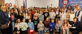 Grupowe zdjęcie dzieci i dorosłych wykonane w sali konferencyjnej. Uczestnicy stoją i klęczą w kilku rzędach. W tle widoczne są banery Wojewódzkiej Stacji Sanitarno‑Epidemiologicznej w Białymstoku oraz flagi. Część dzieci trzyma materiały informacyjne.