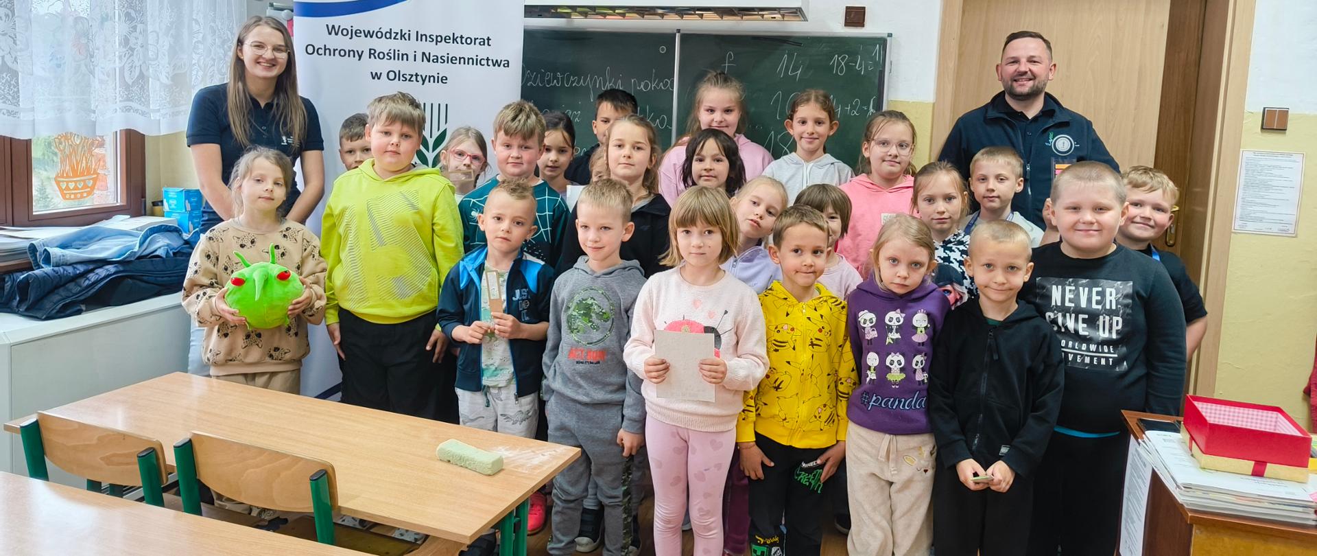 Uśmiechnięte dzieci pozujące do wspólnego zdjęcia w klasie. W tle znajduje się baner z logo PIORiN. Jedna z dziewczynek trzyma w dłoniach zieloną maskotkę PESTY.