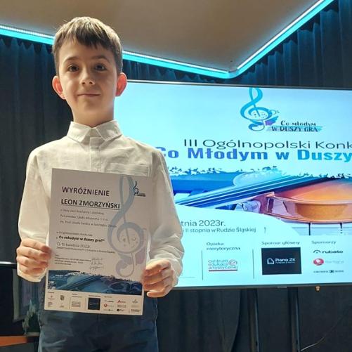 Chłopiec stoi trzymając przed sobą dyplom wyróżnienia, w tle baner z plakatem III Ogólnopolskiego Konkursu "Co młodym d duszy gra?"