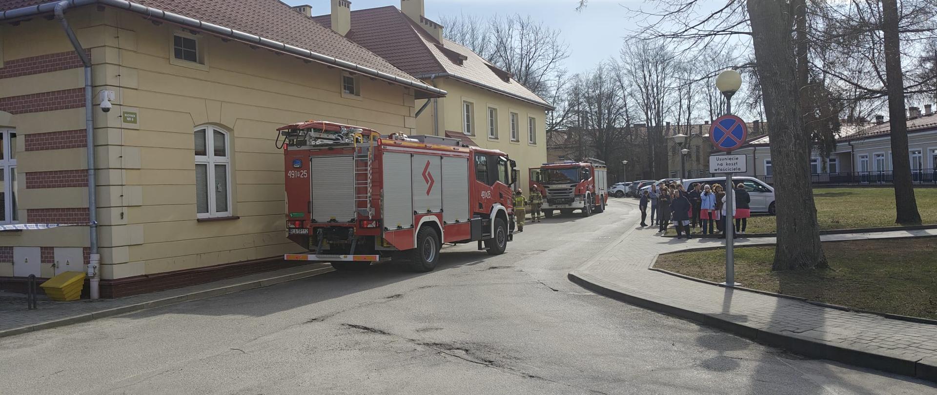 Zdjęcie przedstawia dwa samochody ratowniczo-gaśnicze na terenie centrum medycznego podczas ćwiczeń. Po prawej stoi personel centrym medycznego, który został ewakuowany z budynku.