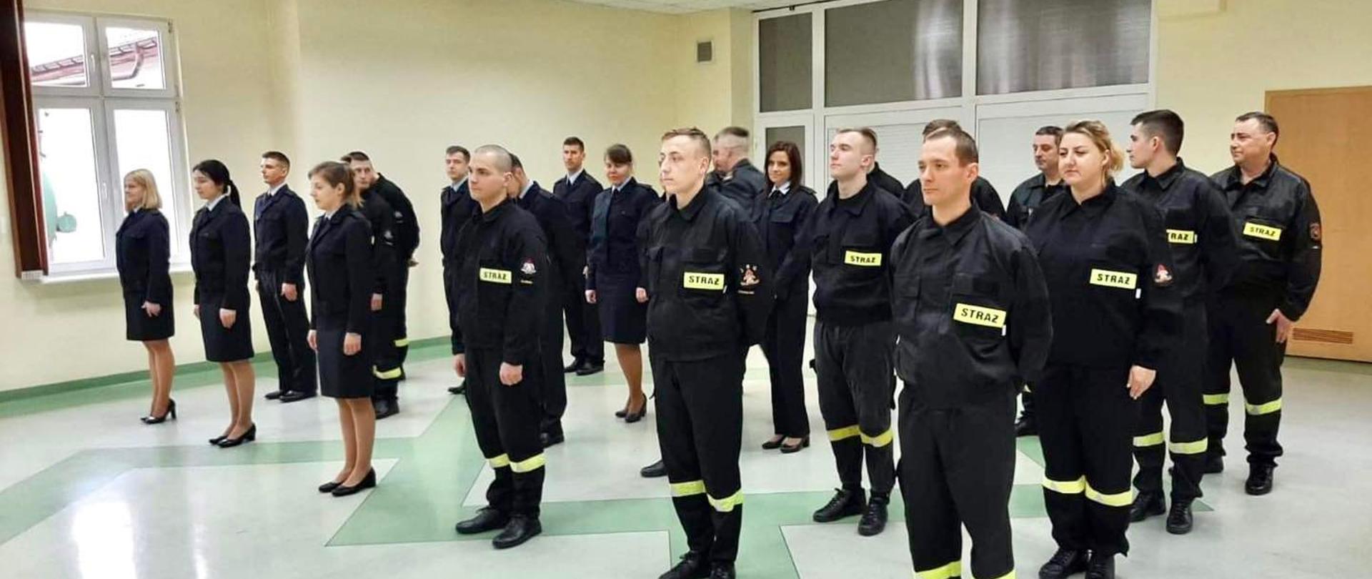 Na zdjęciu widoczni kursantki oraz kursanci szkolenia podstawowego w zawodzie strażak, w ubraniach koszarowych oraz służbowych.
